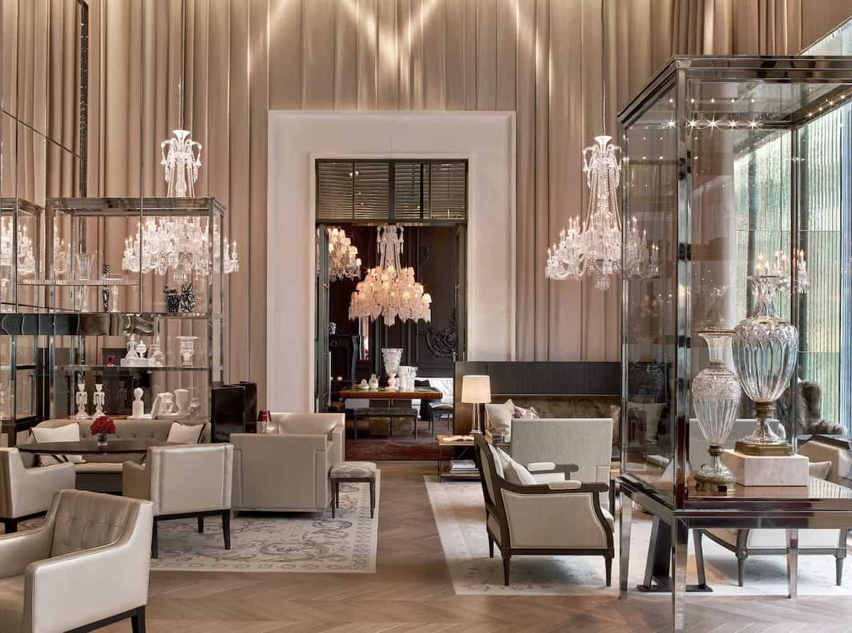 Opulent lounge interior at the Baccarat Hotel New York featuring high ceilings with floor-to-ceiling pleated drapes, sparkling crystal chandeliers, and luxury neutral-toned armchairs. Glass display cases showcase fine crystal glassware alongside polished wood floors and a grand doorway leading to a dark, elegant dining room.