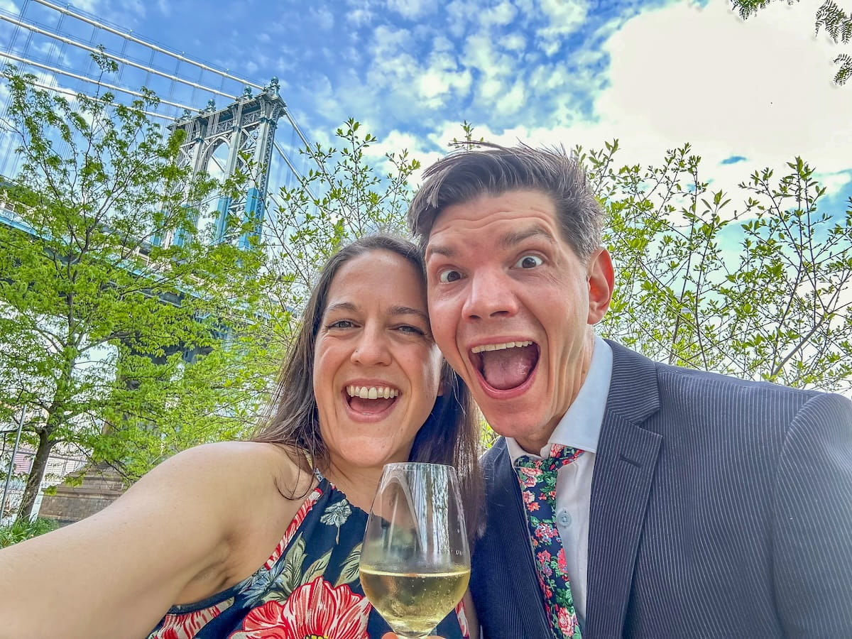 A smiling couple sitting at a waterfront restaurant table in DUMBO, Brooklyn, celebrating their anniversary in NYC with the Manhattan Bridge visible in the background.